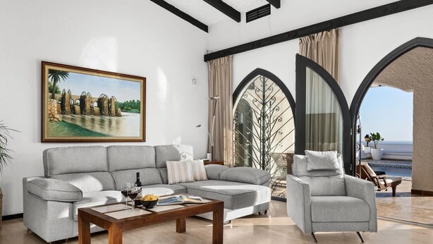 Een moderne woonkamer met een grijze bank, een houten salontafel en een grijze fauteuil. Aan de muur hangt een schilderij, er is een balkon met uitzicht en een groot raam met gordijnen.
