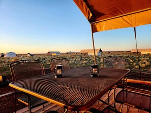 A dining table with chairs under a tent, set in a field with yurts and a clear sky.