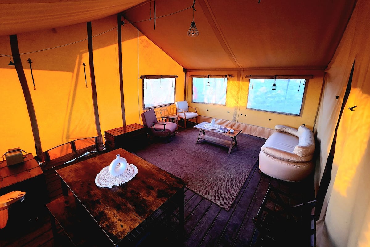 A tented accommodation with a wooden table, a cushioned chair, a sofa, and a small rug.