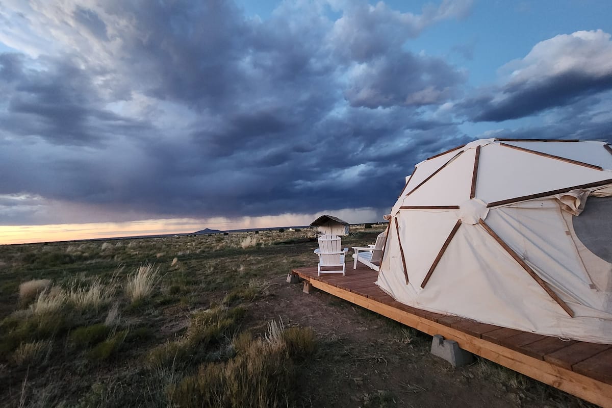 A large tent with a wooden deck and two white chairs, set in a grassy field with a cloudy sky.