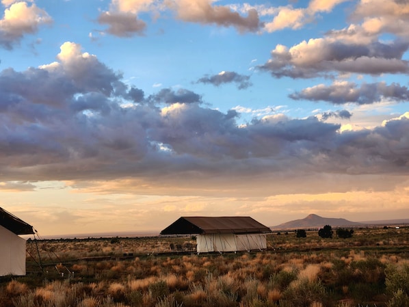 A rustic outdoor setting with a large tent-like structure, a smaller white building, and a mountainous backdrop under a partly cloudy sky.
