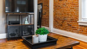 A living room with a brick wall, a flat-screen TV, a wooden coffee table, and a potted plant. - Cozy, Historic Home in A++ Location!! (Denver)