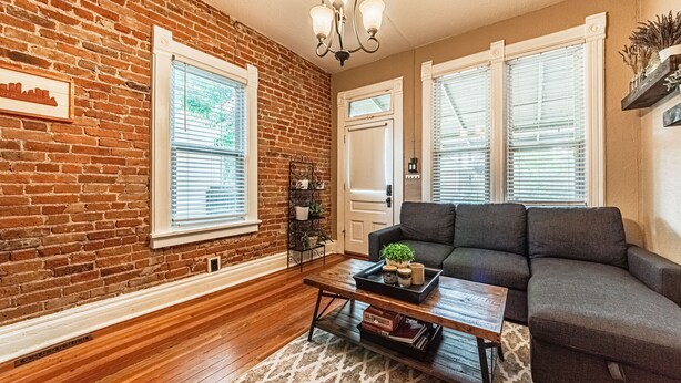 A living room with a brick wall, wooden flooring, a gray sofa, a coffee table with decorative items, and a chandelier. - Cozy, Historic Home in A++ Location!! (Denver)