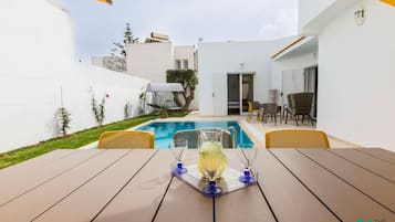 A poolside area with a table set for two, a covered seating area, and a view of the pool and adjacent buildings.