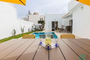 Zona de piscina con mesa para dos, un área de estar cubierta y vista a la piscina y edificios cercanos.