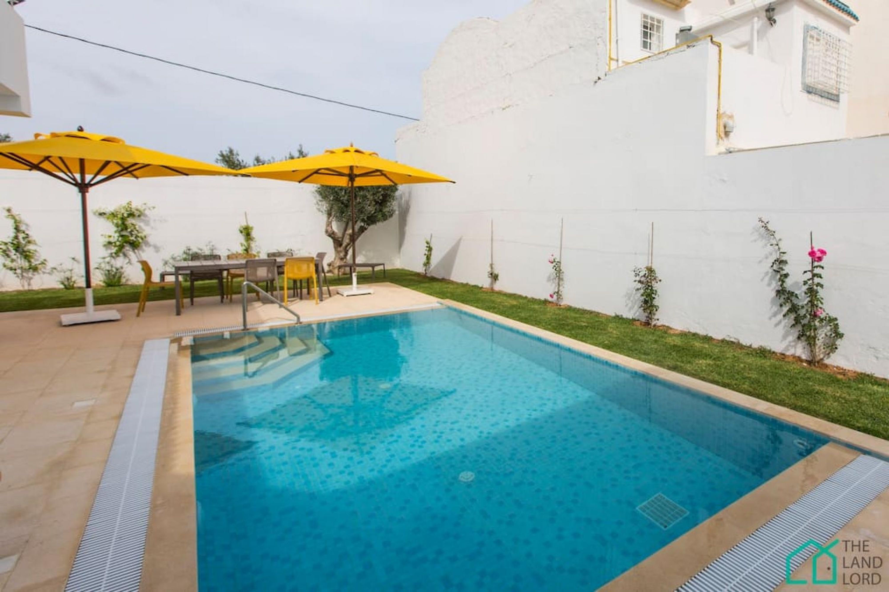 A swimming pool with yellow umbrellas, a table, chairs, and a small tree.