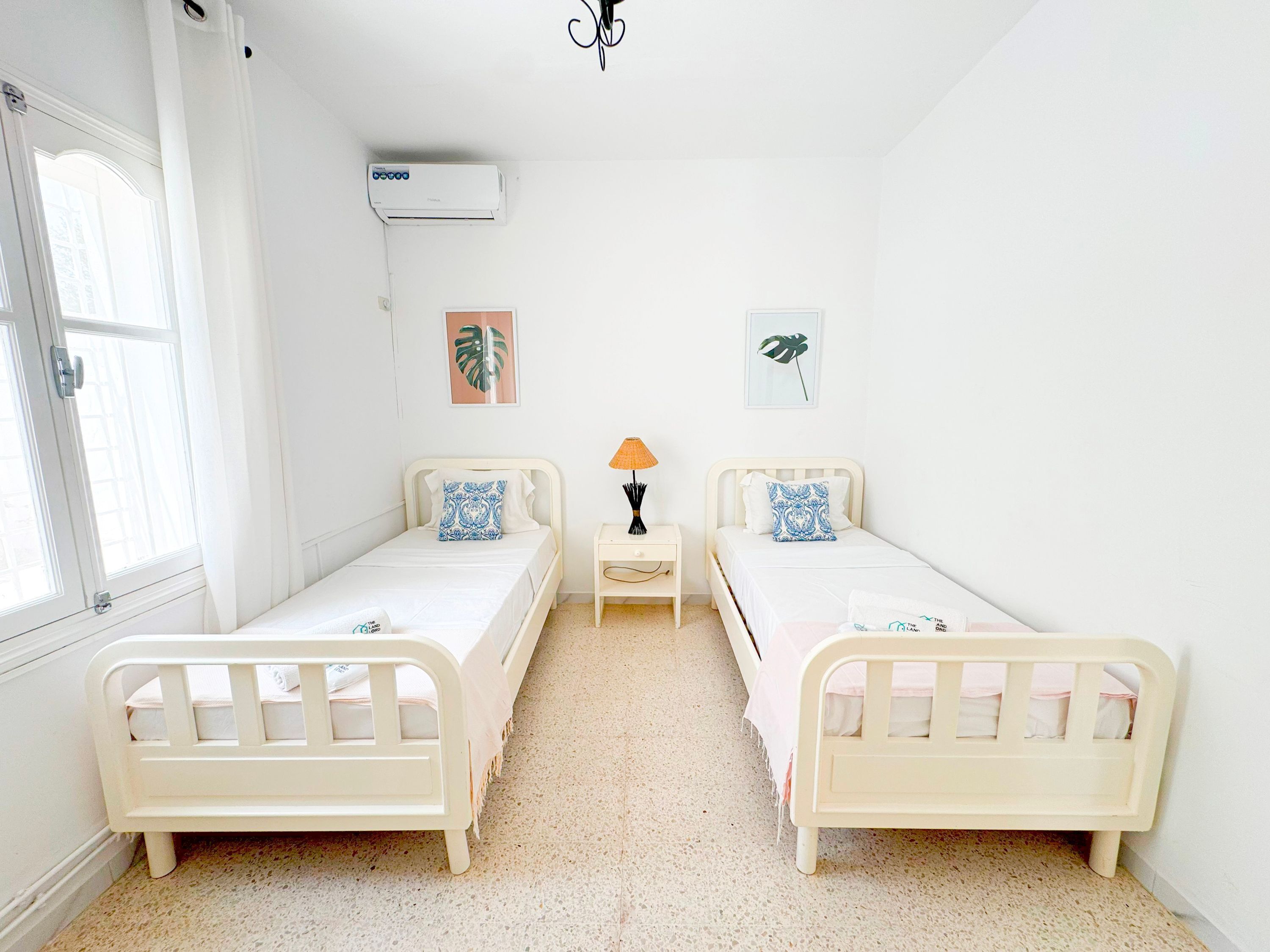 A room with two single beds, a nightstand with a lamp, and an air conditioning unit on the wall.