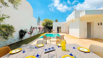 An outdoor dining area with a pool, white walls, and a clear blue sky.