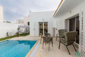 Un área de piscina con una piscina azul cristalina, una zona de estar techada con sillas, y un ventanal corredizo que da a un patio.