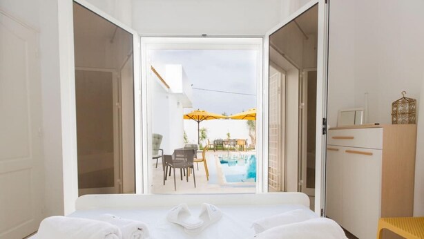 Hotelkamer met een bed, twee opgerolde handdoeken, een houten kast en uitzicht op een buitenzwembad met stoelen en parasols.