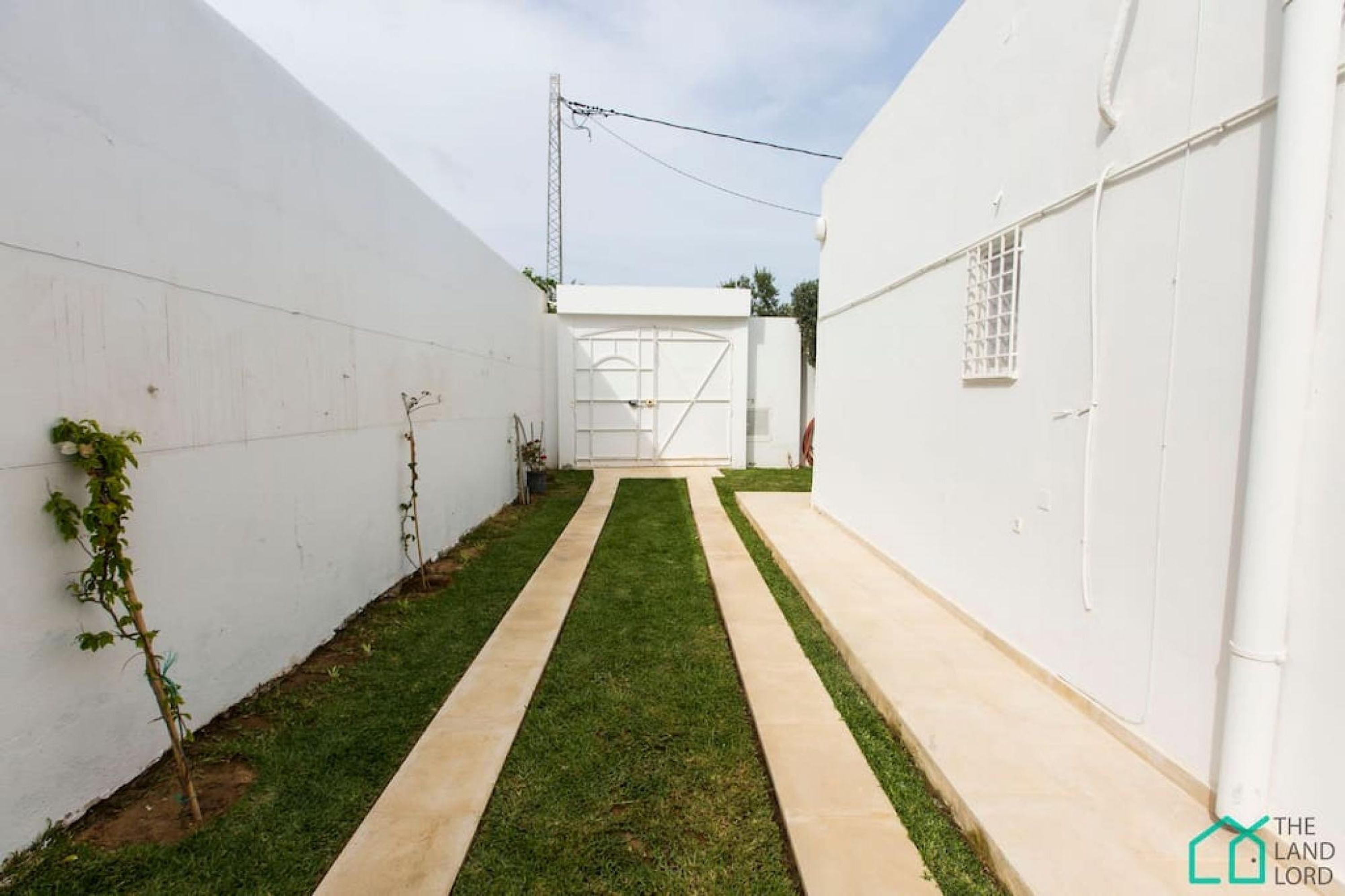 A narrow pathway flanked by white walls, leading to a gated entrance.