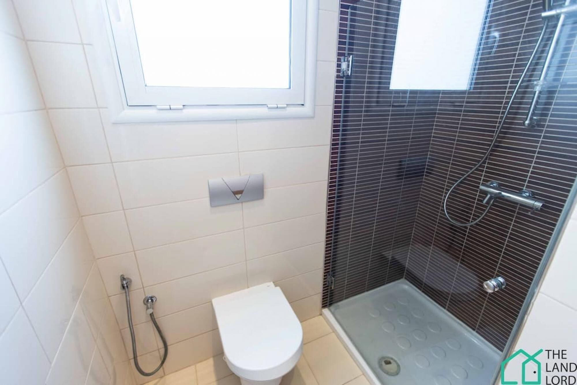 Une petite salle de bain moderne, équipée de toilettes, d’une douche avec une porte en verre et d’une fenêtre située au-dessus des toilettes.