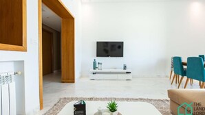 A modern living room with a flat-screen TV mounted on the wall, a white console table, teal chairs, and a beige sofa.
