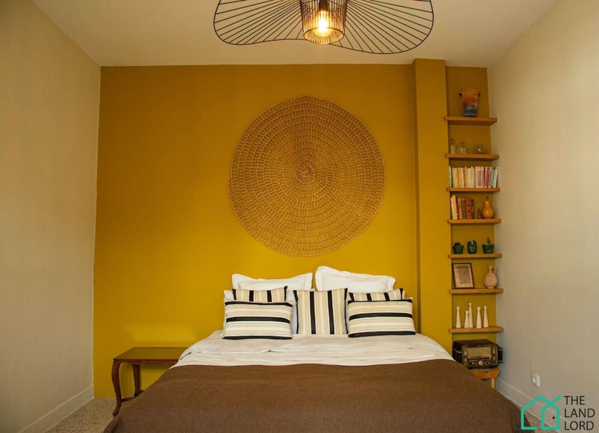 A bedroom with a bed, a circular wall decoration, a shelf with books and decorative items, and a small table.