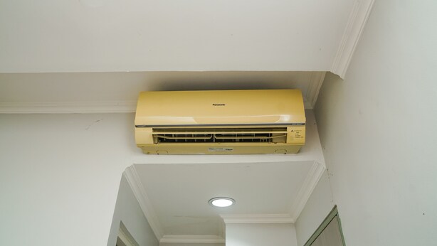A yellow Panasonic air conditioner mounted on a white ceiling in a narrow hallway. - Singgahsini Rock Gubeng (Surabaya)