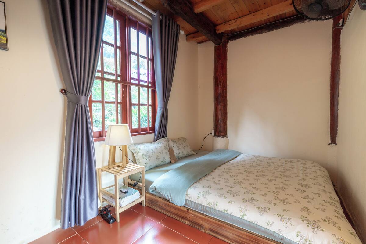 Quarto com cama, mesa de cabeceira, abajur e janela com cortinas.