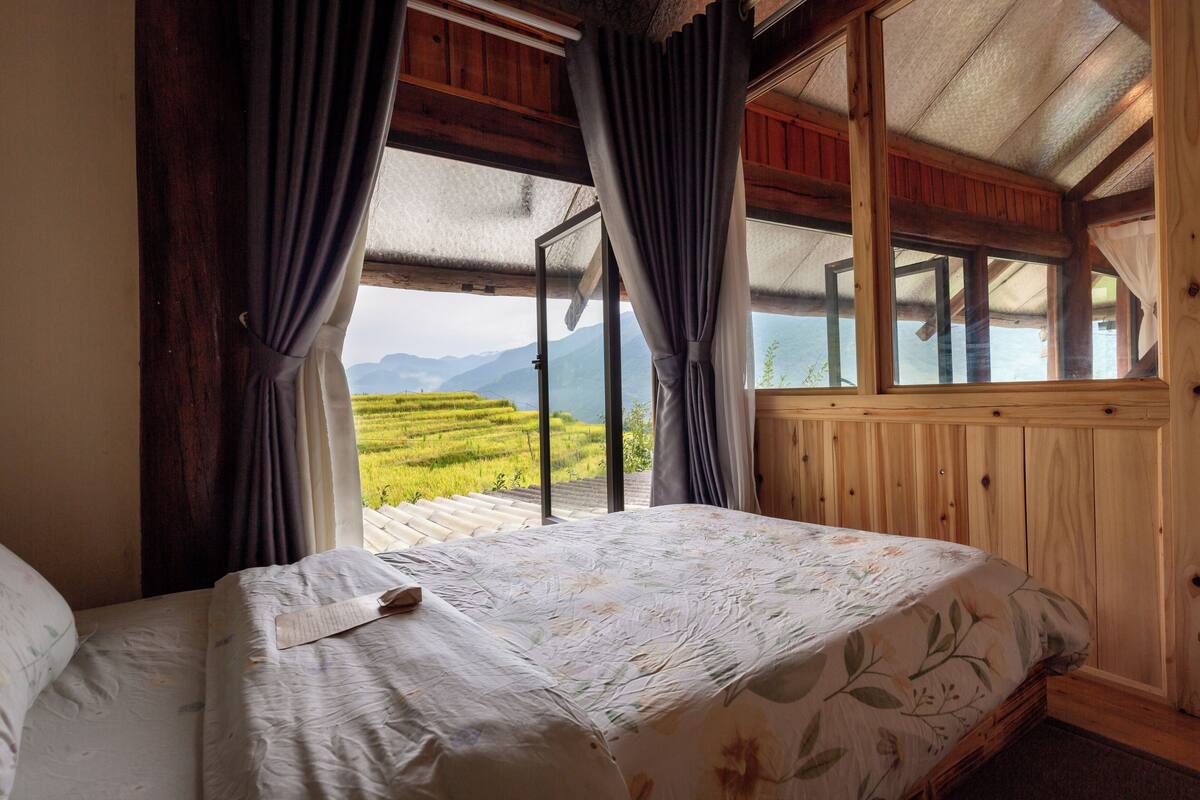 Quarto com cama, paredes de madeira e uma janela grande com vista para uma paisagem verde.