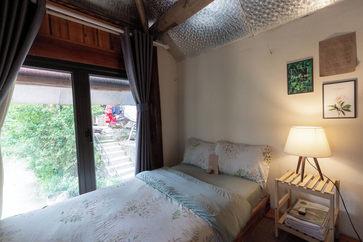Quarto com cama, mesa de cabeceira com abajur, janela com cortinas e vista para o exterior.