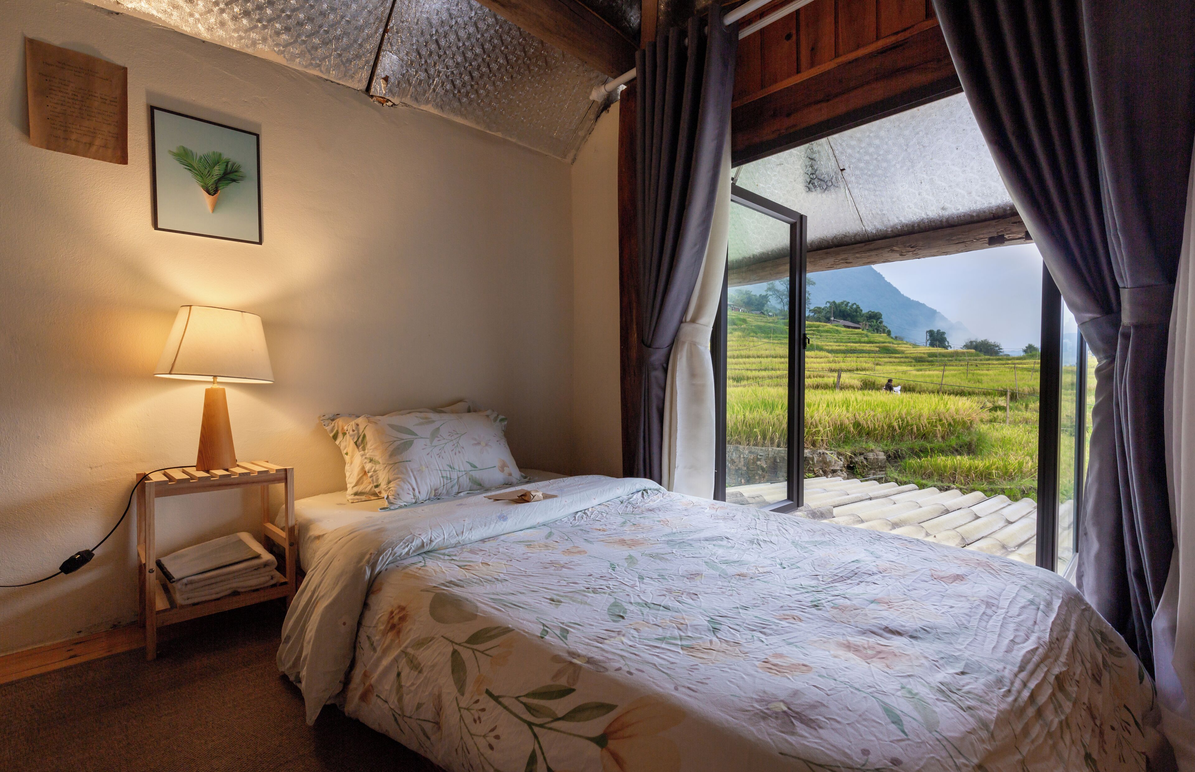 Quarto com cama, luminária de cabeceira, quadro emoldurado e janela com cortinas.