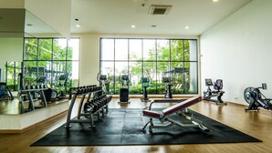 Ein Fitnessstudio mit Cardiogeräten, Kurzhanteln und einer Langhantelbank. Große Fenster bieten Blick auf die grüne Umgebung.