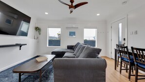 Un salon moderne avec un canapé d’angle, une table à manger avec des chaises, un ventilateur de plafond et de grandes fenêtres.