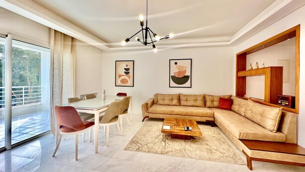 Una sala de estar moderna con un sofá modular, una mesa de comedor y una mesa de centro. Hay pinturas abstractas en las paredes y un ventanal que ofrece vistas al exterior.