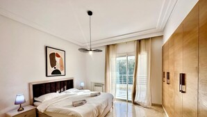Un dormitorio moderno con una cama grande, una mesita de noche con lámpara, una luz de techo, una ventana con cortinas y un armario de madera.