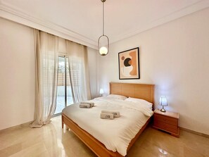 A spacious bedroom with a large bed, a wooden headboard, a bedside table with a lamp, and a view through a sliding glass door.