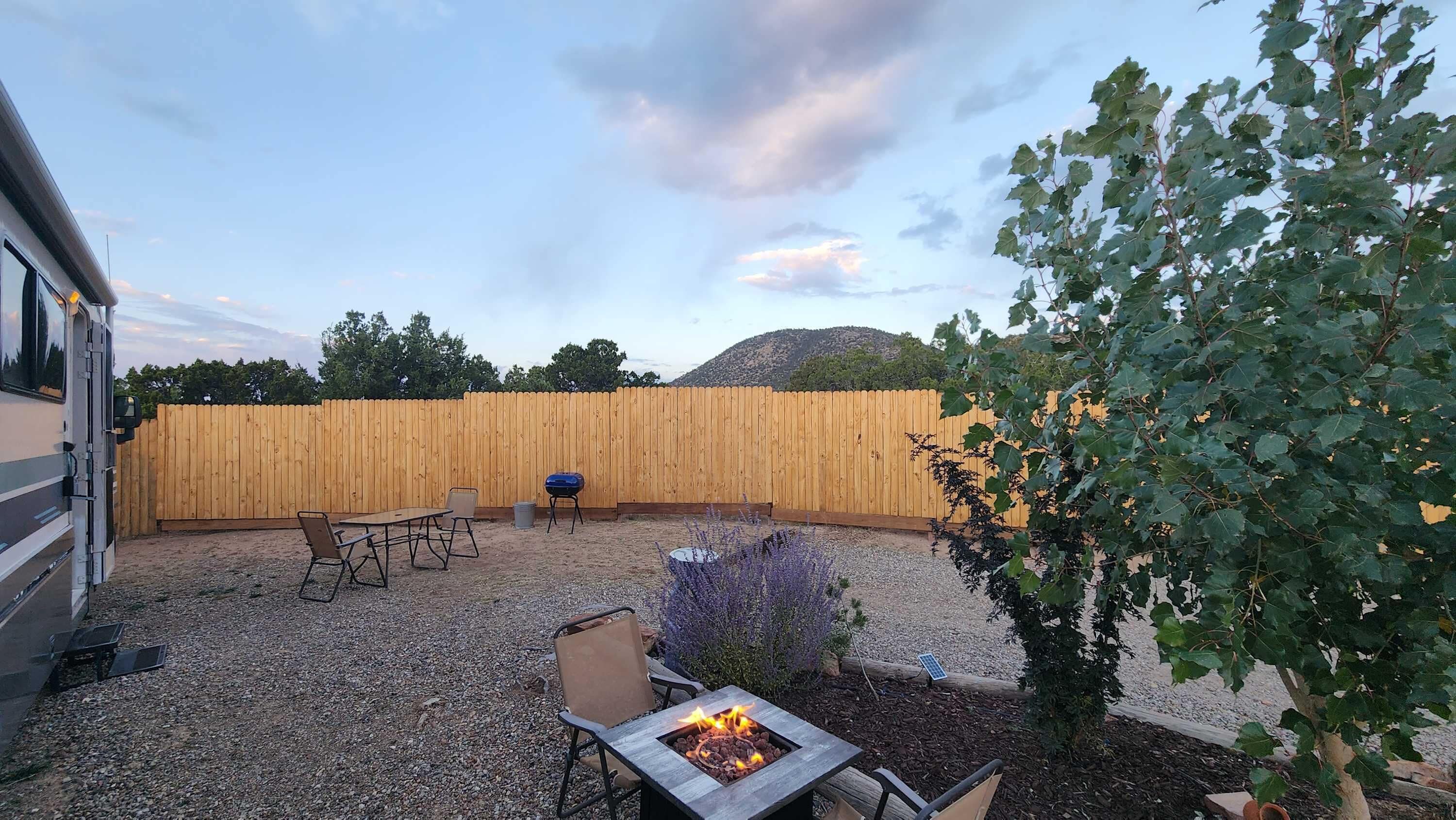 A backyard with a wooden fence, a fire pit, outdoor furniture, and a tree.