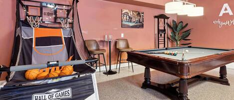 Una mesa de billar en una habitación con mesa de billar, aro de baloncesto y una bolsa con el texto 'Hall of Games'.
