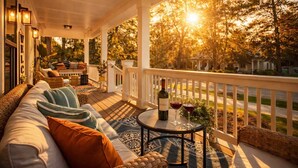 A covered porch with a sofa, a table with a bottle and glasses, and a view of trees and houses.