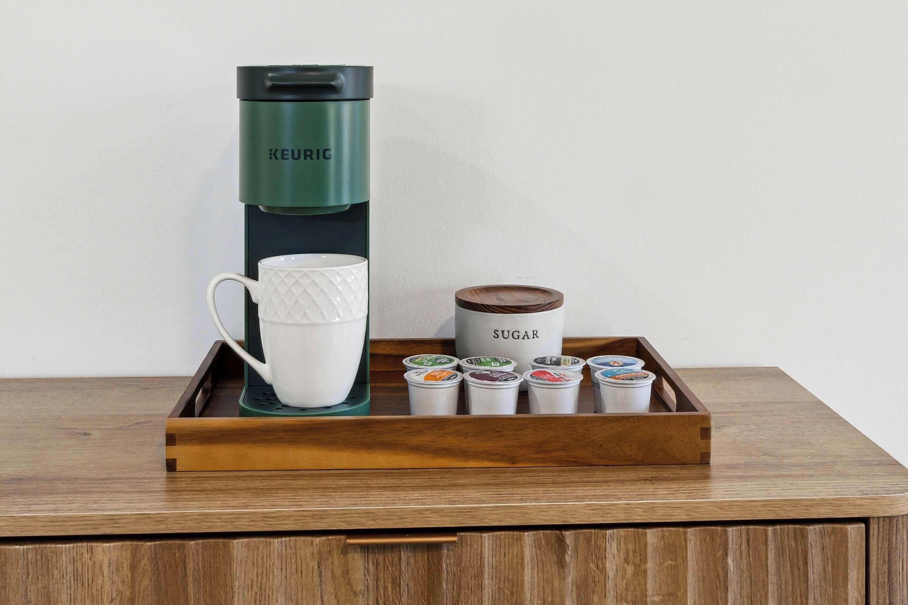 Una cafetera Keurig, una taza blanca, un recipiente para azúcar y varios sobre de azúcar sobre una bandeja de madera.