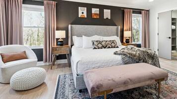 A modern bedroom with a large bed, bedside lamps, a chair, a bench, and artwork on the wall.