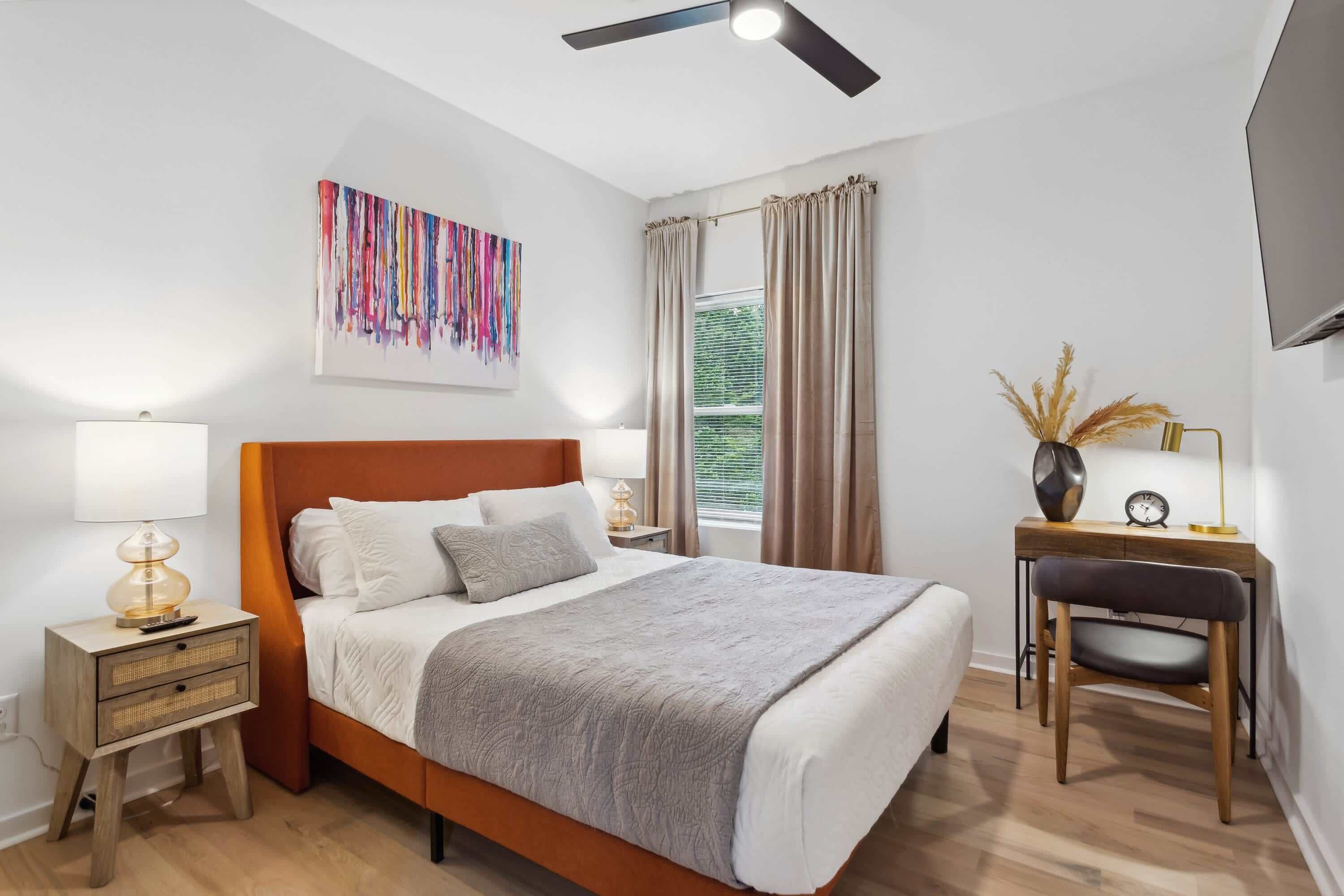 A modern bedroom with a wooden bed, bedside lamps, a desk with a chair, and a ceiling fan.