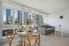 A modern living room with a dining area, large windows, a grey sofa, and a view of high-rise buildings.
