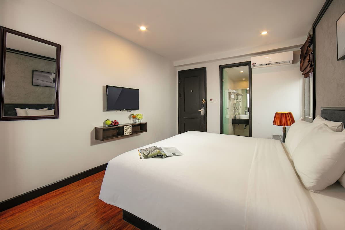 A modern hotel room with a large bed, a flat-screen TV, a small shelf with decorative items, and a door leading to a bathroom.