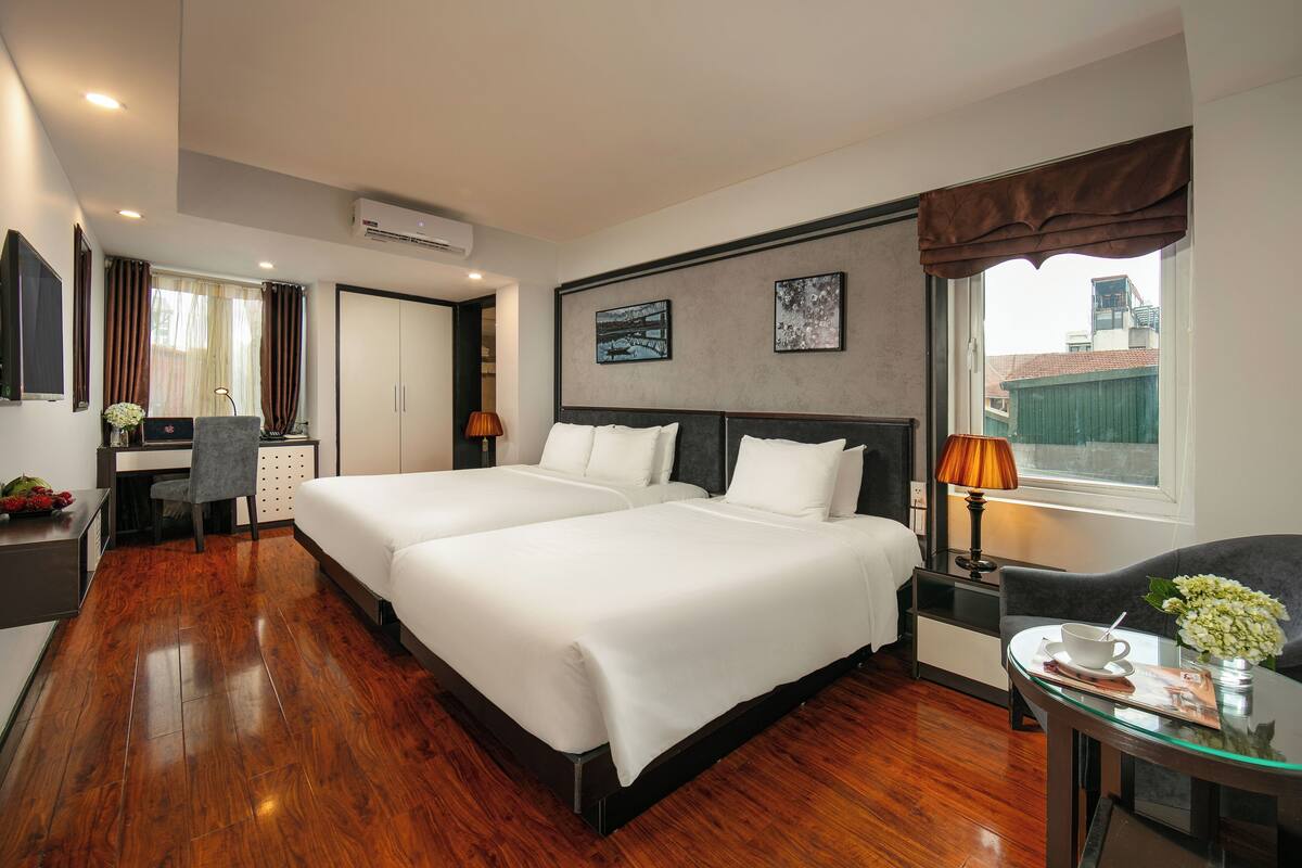 A hotel room with two beds, a desk, a chair, a lamp, and a window with curtains.
