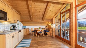 Interior de madera con cocina, comedor y grandes ventanales con vistas a la montaña.