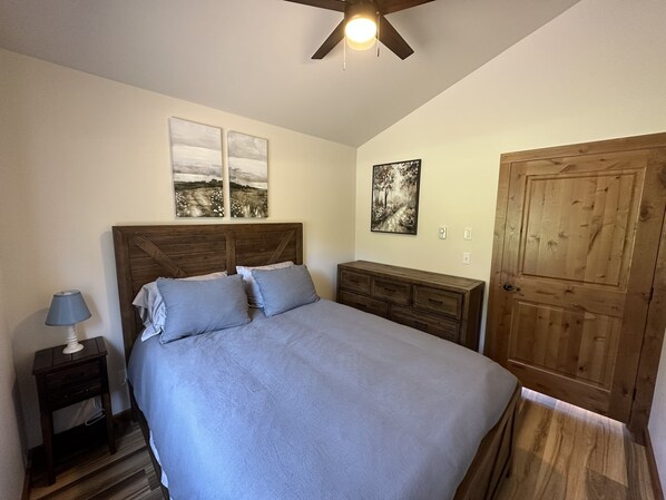 A bedroom with a wooden headboard, a bed with grey bedding, a nightstand with a lamp, a dresser, and a wooden door. - Cozy guest cabin (Kalispell)