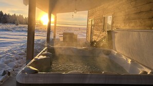Eine Holzhütte mit Whirlpool im Vordergrund, einer Holzterrasse und Blick auf eine verschneite Landschaft, in deren Hintergrund die Sonne untergeht.