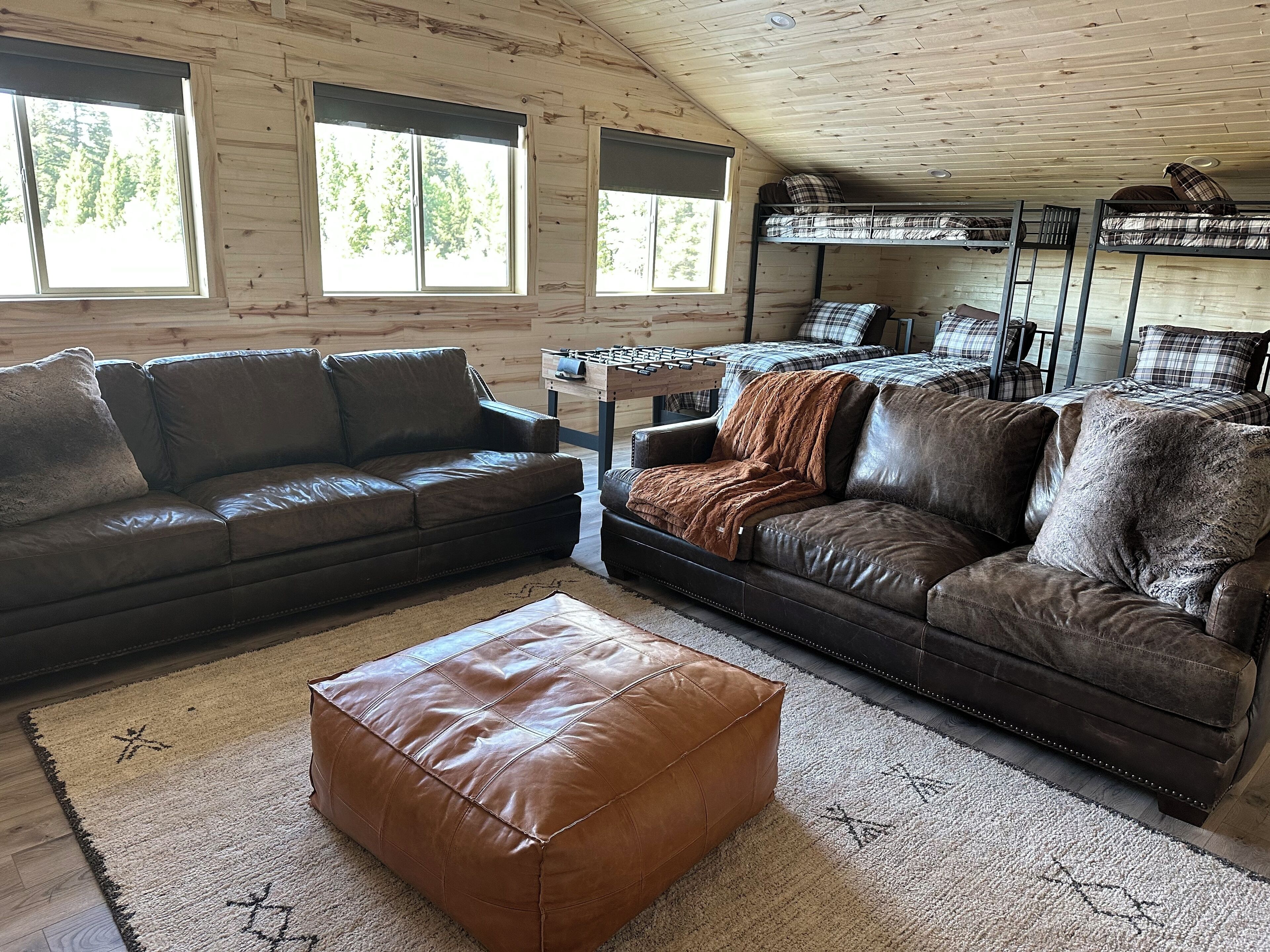A cozy cabin interior with two leather sofas, a brown ottoman, and a wooden ceiling.