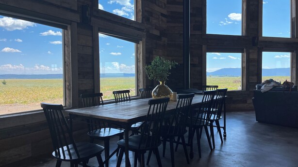 Ein Essbereich mit großem Tisch und Stühlen, großen Fenstern mit Blick auf ein Feld und Berge, und einer Topfpflanze auf dem Tisch.