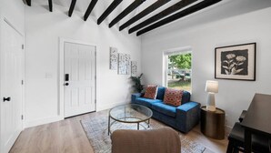 Uma sala de estar moderna com um sofá azul, uma mesa de centro de vidro e uma cadeira de vime. Há uma porta, uma janela e um abajur sobre uma mesa lateral.