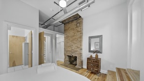 A modern bedroom with a brick fireplace, wooden flooring, and a white bed. - Huge Retreat| Rooftop Deck| Stay At Findlay Market (Cincinnati)