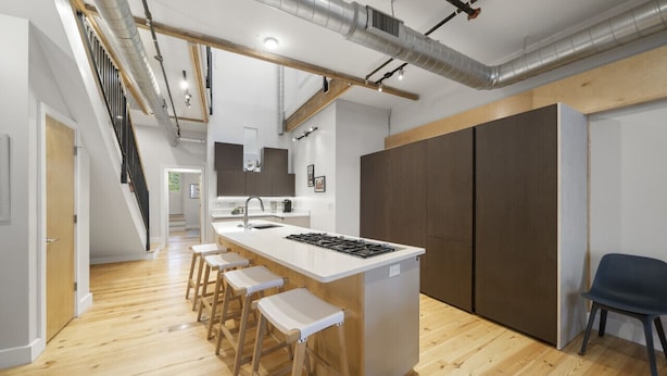 A modern kitchen with a central island, wooden stools, a staircase, and a large wardrobe. - Huge Retreat| Rooftop Deck| Stay At Findlay Market (Cincinnati)