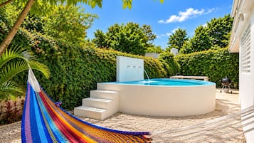 A swimming pool with a hot tub, surrounded by a hedge, a hammock, and a barbecue grill.