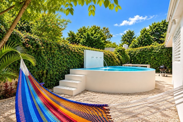 A swimming pool with a hot tub, surrounded by a hedge, a hammock, and a barbecue grill.