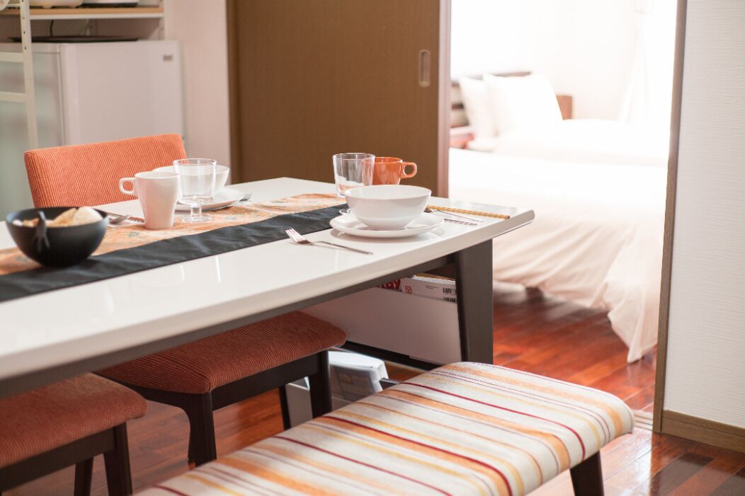 Un comedor con una mesa lista para dos comensales, un banco y una cama al fondo.