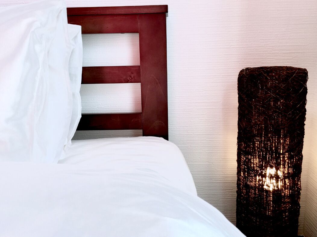 A neatly made bed with white linens and a wooden headboard. A tall, textured lamp stands beside the bed.