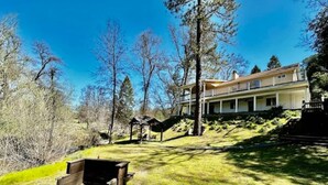 A large house with a covered porch, a wooden deck, and a grassy yard with trees. - Creekside Dream - Dog Friendly/Game Room/Sleeps 12 (Oakhurst)